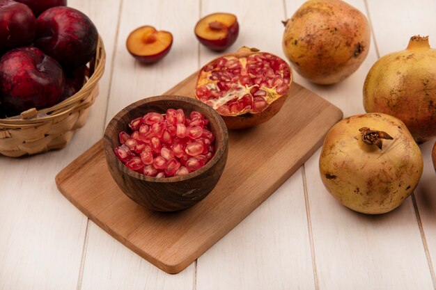 Top view of juicy pomegranate seeds on a wooden bowl on a wooden kitchen board with pluots on a bucket with pomegranates isolated on a white wooden background