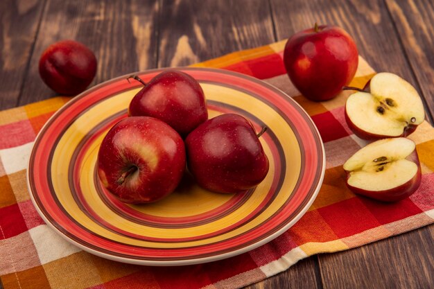 Top view of juicy red apples on a plate on a checked cloth with halved apples isolated on a wooden surface