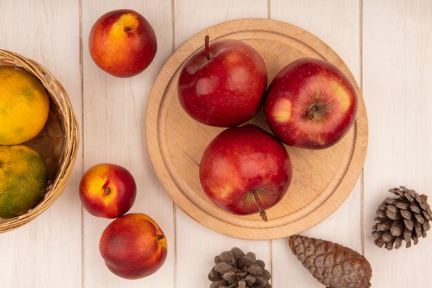 Top view of juicy red apples on a wooden kitchen board with tangerines on a bucket with peaches and pine cones isolated on a white wooden wall