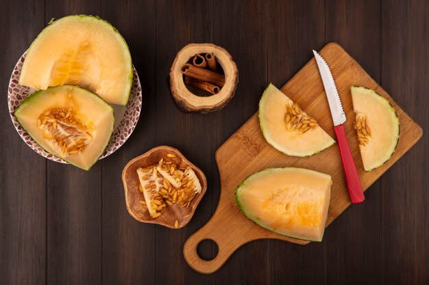 Top view of juicy and sweet slices of cantaloupe melon on a wooden kitchen board with knife with melon seeds on a wooden bowl with cinnamon sticks on a wooden background
