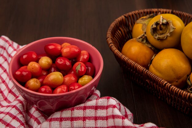 Top view of juicycornelian cherries on a bowl on a red checked cloth with persimmon fruits on a bucket on a wooden surface