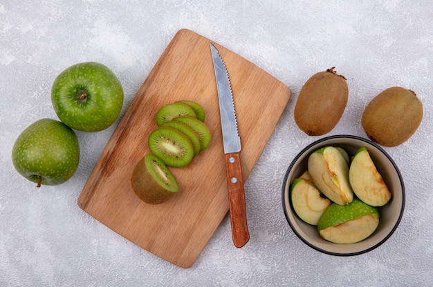 Top view kiwi slices with knife on cutting board with green apple slices in bowl on white background