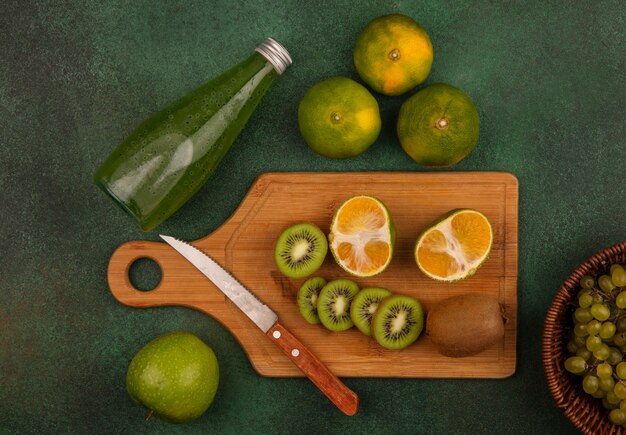 Top view kiwi slices with knife on cutting board with tangerines and bottle of juice on green wall