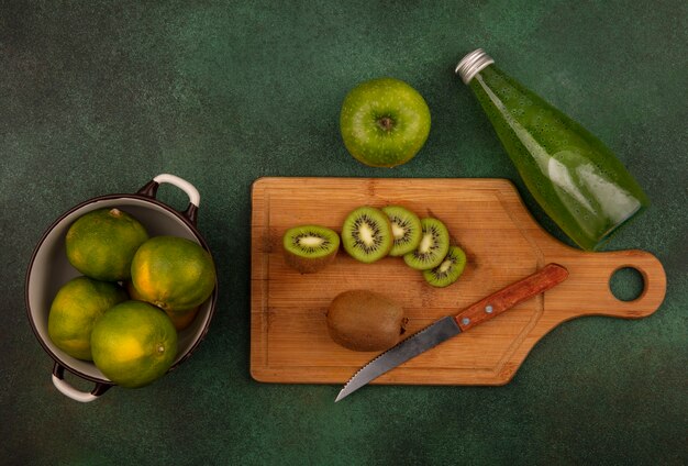 Top view kiwi slices with a knife on a cutting board with tangerines in a saucepan and a bottle of juice on a green wall