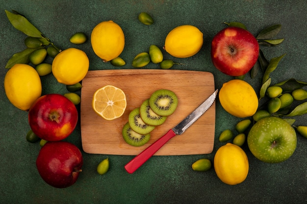 緑の表面に分離されたレモンとカラフルなリンゴとナイフで木製のキッチンボード上のキウイスライスの上面図