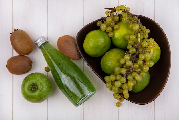 Top view kiwi with bottle of juice apple and grapes with tangerines in bowl on white wall