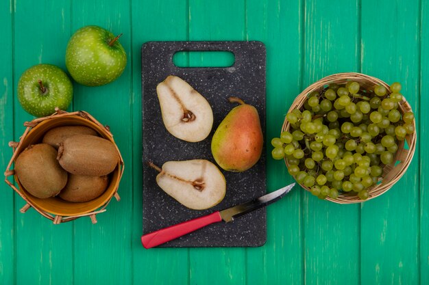 커팅 보드에 칼과 녹색 배경에 녹색 사과 바구니와 배 조각에 녹색 포도와 상위 뷰 키위