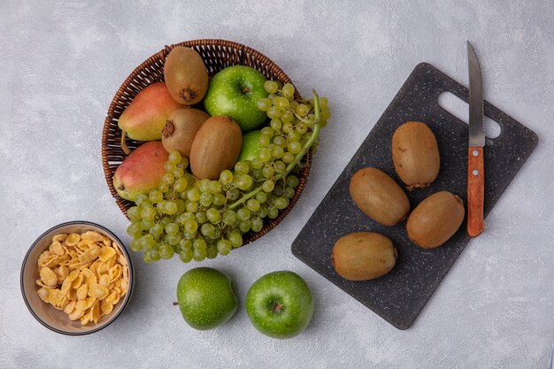 Top view kiwi with a knife on a cutting board  with green apples  grapes and pear in a basket  with cornflakes in a bowl on a white background