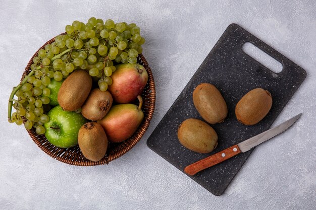 Top view kiwi with a knife on a cutting board with green apples  grapes and pears in a basket on a white background