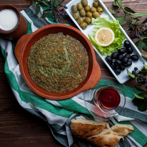 Top view kuku with glass of tea and assorted olives and bread in clay plate