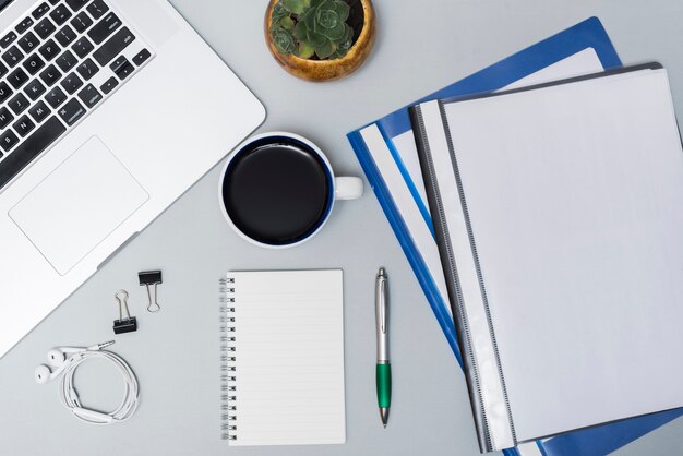 Top view of laptop; folders; coffee cup; earphone; spiral notepad and pen against gray background