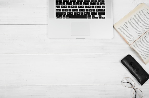 Top view of laptop with book and eyeglasses on white wooden table