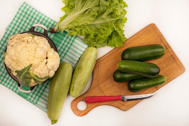 Top view of large white cauliflower on a bowl on a green checked cloth with cucumbers on a wooden kitchen board with knife with zucchinis and lettuce isolated on a white surface