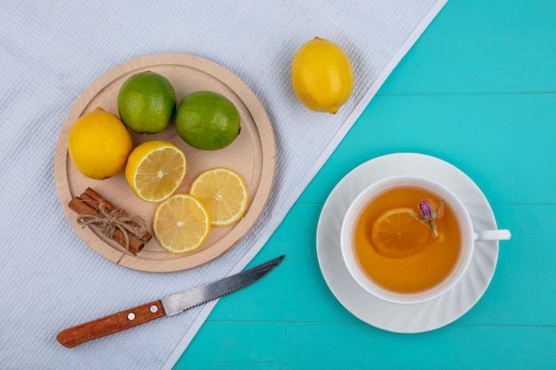 Top view lemon with lime on a tray with cinnamon a knife and a cup of tea on a white towel on a light blue background