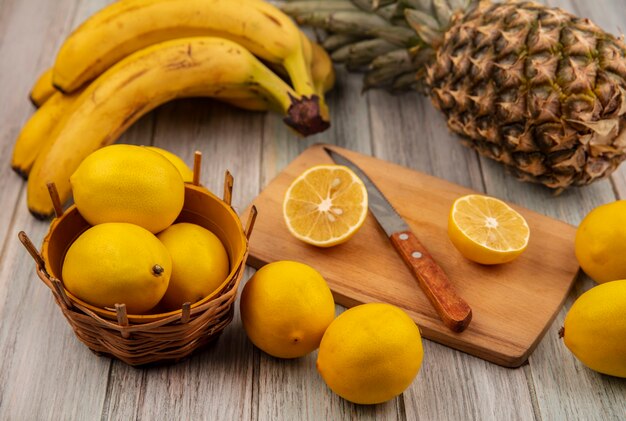 Top view of lemons on a bucket with half lemon on a wooden kitchen board with knife with lemons bananas and pineapple isolated on a grey wooden surface