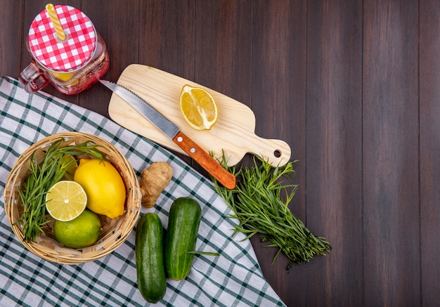 Top view of lemons on a bucket with tarragon green on a checked tablecloth with cucumbers on a wooden surface