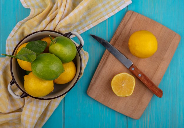 まな板の上にナイフとターコイズブルーの背景に黄色の市松模様のタオルと鍋にライムとライムの上面図レモン