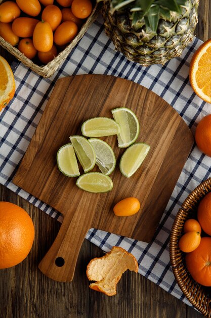 Вид сверху на ломтики лайма и кумкват на разделочной доске с кумкватом, ананасом, мандарином, нарезанным апельсином на клетчатой ткани на деревянном фоне