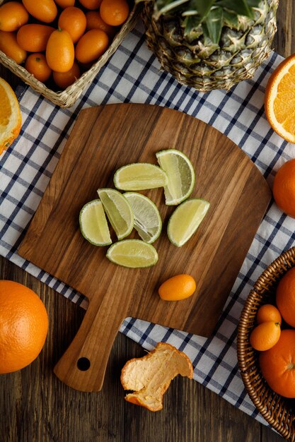 Top view of lime slices and kumquat on cutting board with kumquats pineapple tangerine cut orange on plaid cloth on wooden background