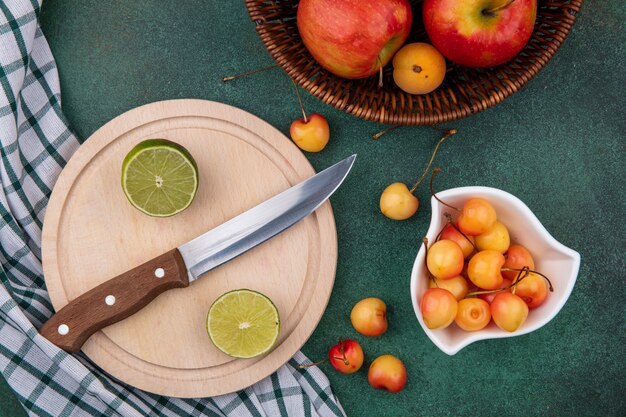 Top view of lime slices with a knife on a stand with white cherries and apples in a basket on a green surface