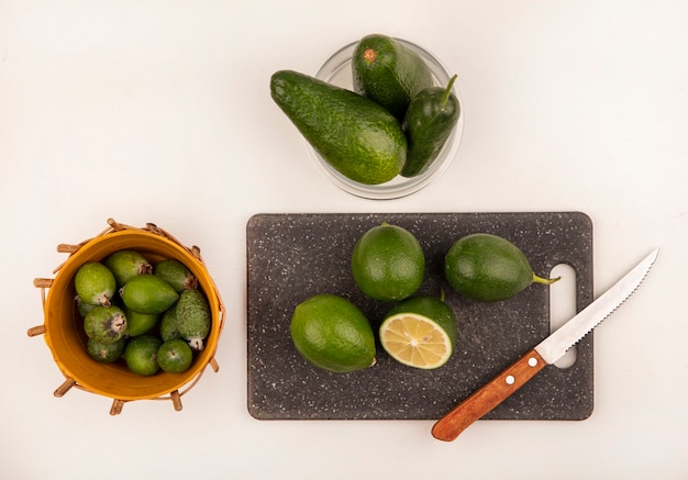 Top view of limes on a kitchen board with knife with avocados and cucumber on a glass bowl with feijoas on a bucket on a white wall