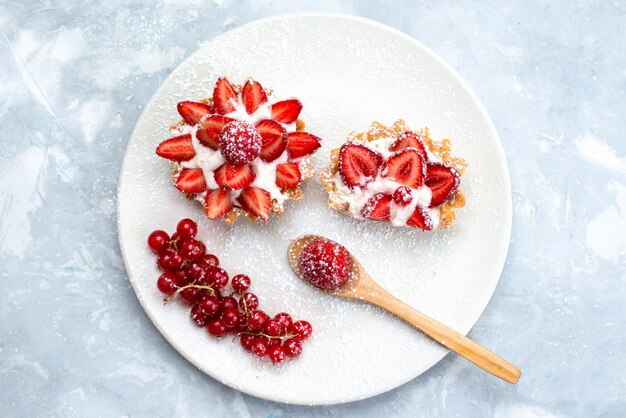 A top view little cake with cream and sliced fresh strawberries inside white plate along with fresh red cranberries on the grey-blue desk