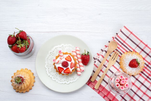 白い机の上のクリームとスライスしたイチゴケーキキャンディー、フルーツケーキベリー甘い砂糖と小さなケーキの上面図