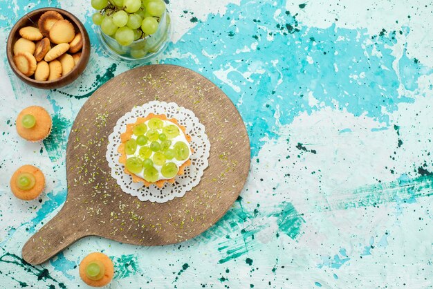 Top view of little cake with delicious cream and sliced and fresh grapes cookies isolated on blue light desk, cake sweet fruit