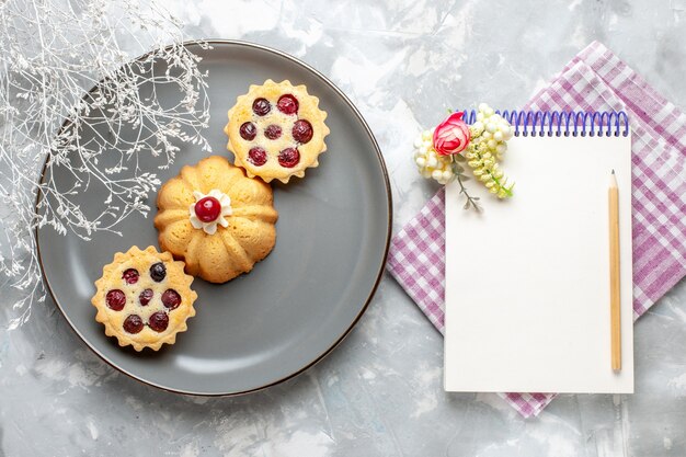 Top view little cakes inside grey plate with notepad on the light background cake sweet sugar bake fruit