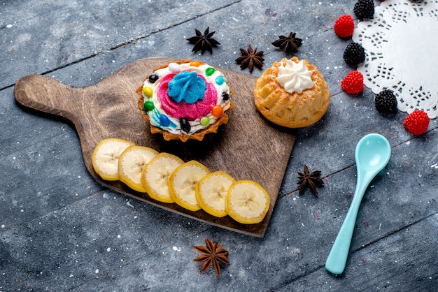 Вид сверху маленькие пирожные со сливками вместе с нарезанными фруктами ягодами синей ложкой на светлом столе торт бисквитный сладкий сахар фрукты