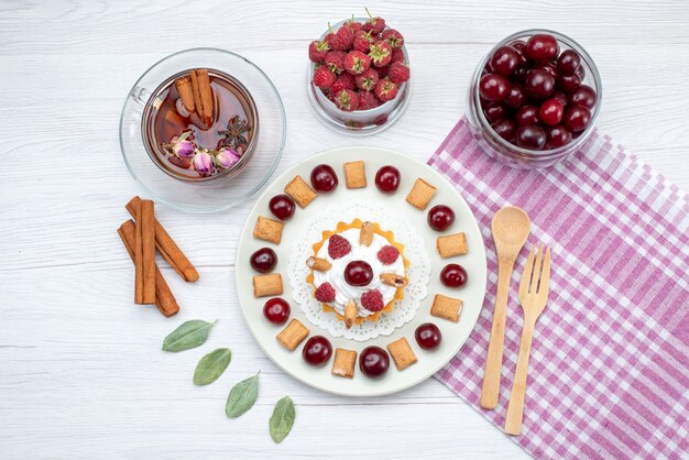 Top view little creamy cake with raspberries cherries and little biscuits tea cinnamon on the white-light desk fruit cake sweet berry cream