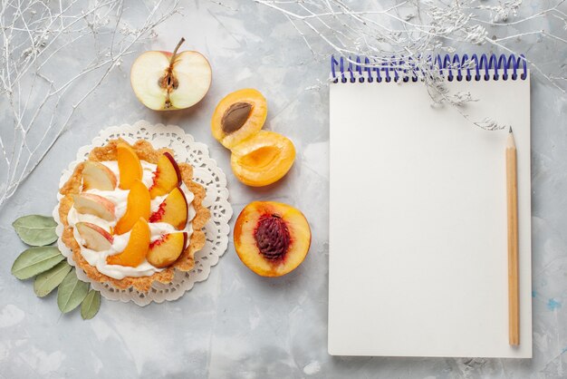 Вид сверху маленький сливочный торт с нарезанными фруктами и белым кремом вместе со свежими абрикосами и персиками на белом столе фруктовый торт бисквитное печенье