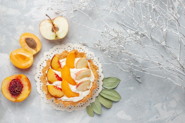 Вид сверху маленький сливочный торт с нарезанными фруктами и белым кремом вместе со свежими абрикосами и персиками на белом столе, фруктовый торт, бисквитное печенье, выпечка