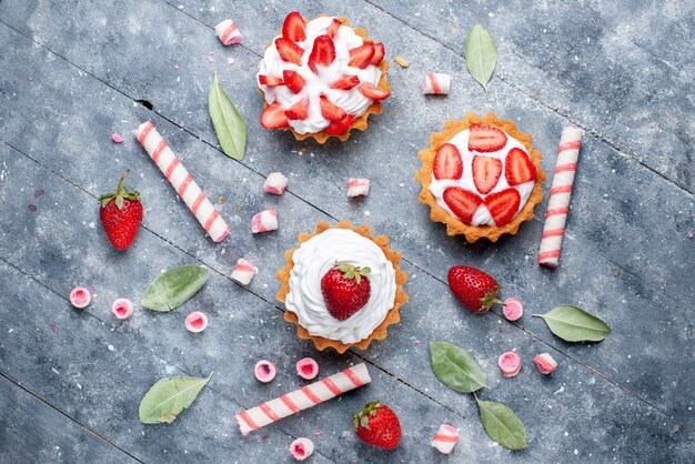 Top view little creamy cakes with sliced and fresh strawberries along with stick candies on the grey surface fruit cake sweet bake berry