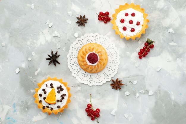 Вид сверху маленькие торты со сливками и разными фруктами, изолированные на светлой поверхности, сахар, сладкий чай