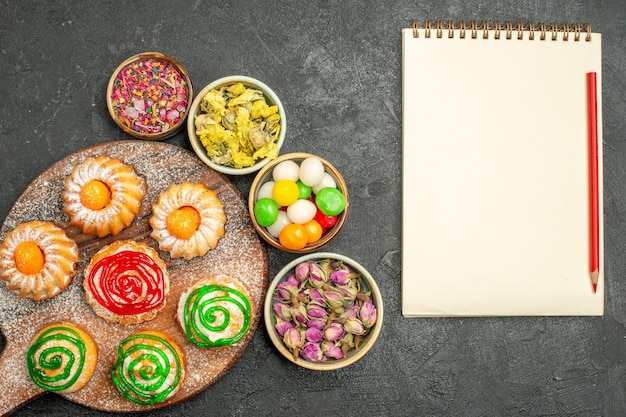 Top view of little delicious cakes with candies and flowers on dark