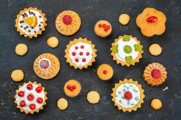 Top view little delicious cakes with cream and fresh fruits on the dark surface sweet biscuits cake sugar