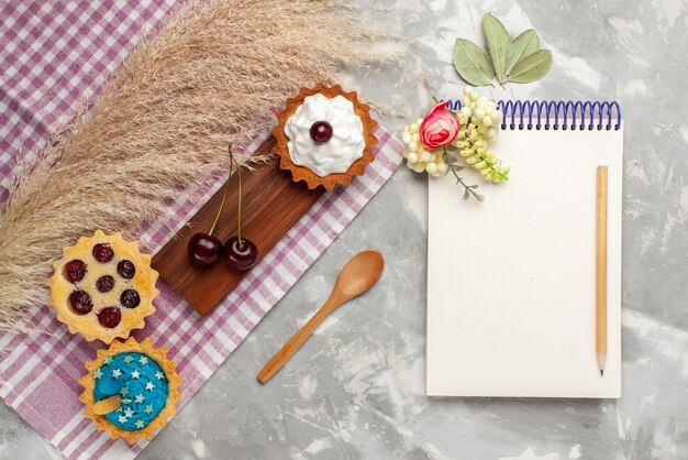 Top view little delicious cakes with cream and fruits notepad on light-white background cake sweet cream bake fruit