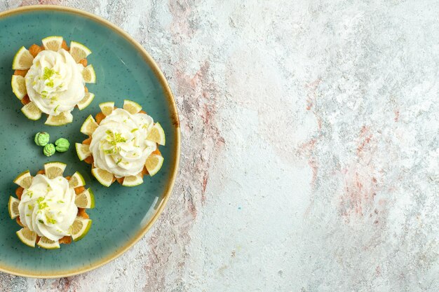 Вид сверху маленькие вкусные пирожные со сливками и дольками лимона на белом письменном столе, печенье, печенье, сладкий чай, сахар