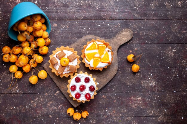 Top view little delicious cakes with cream sliced fruits and fresh yellow cherries on the brown wooden background fresh fruit cake biscuit sweet