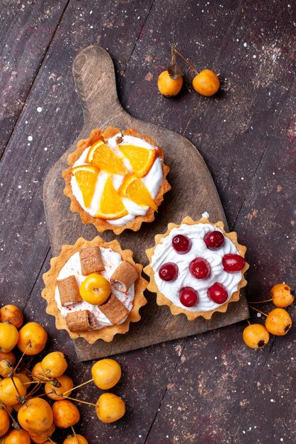 Top view little delicious cakes with cream sliced fruits and fresh yellow cherries on the brown wooden desk fresh fruit cake biscuit sweet