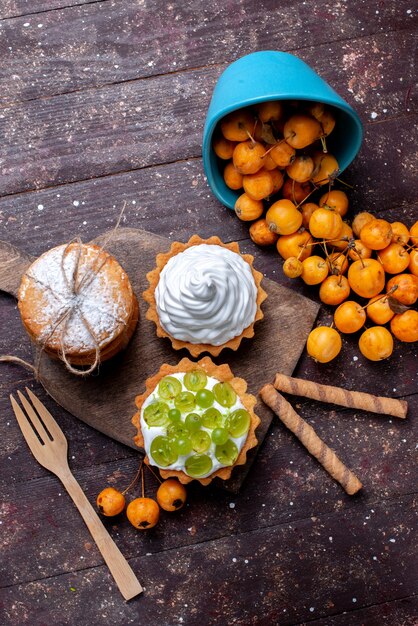 Вид сверху на маленькие вкусные пирожные с кремом, ломтиками виноградного печенья и свежей желтой вишней на коричневом столе, свежий фруктовый торт, печенье, черешня