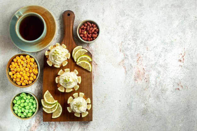 Вид сверху маленькие вкусные пирожные с чашкой чая и конфетами на белой поверхности торт бисквитное печенье сладкий чай крем