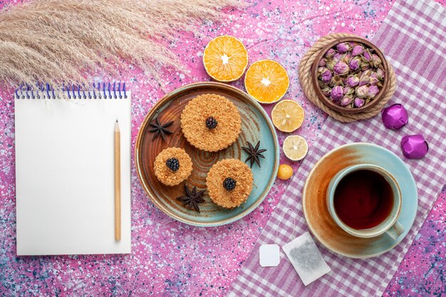 Top view of little delicious cakes with orange slices and tea on the light-pink surface