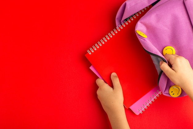 Top view little kid preparing for school taking copybook from bag on red surface