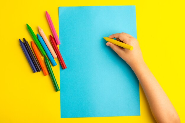 Top view little kid using colorful pencils on blue paper on the yellow surface