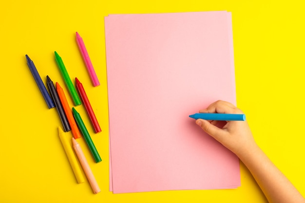 黄色の表面のピンクの紙にカラフルな鉛筆を使用して小さな子供を上面図