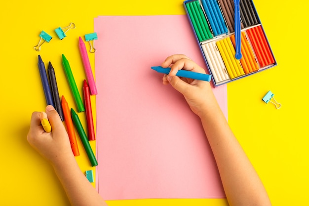 Top view little kid using colorful pencils on pink paper on yellow surface