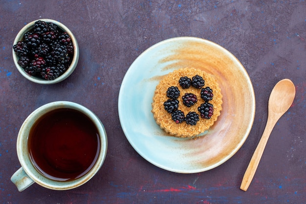 Top view of little round cake with berries inside plate along with cup of tea on dark surface