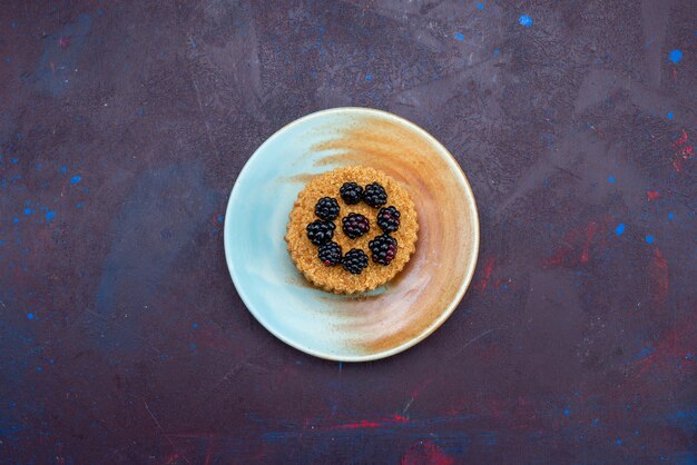 Top view of little round cake with berries inside plate on dark surface